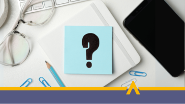 Biurko z notatnikiem, na którym leży niebieska karteczka z dużym znakiem zapytania, obok okulary, ołówek i smartfon.