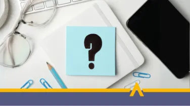 Biurko z notatnikiem, na którym leży niebieska karteczka z dużym znakiem zapytania, obok okulary, ołówek i smartfon.