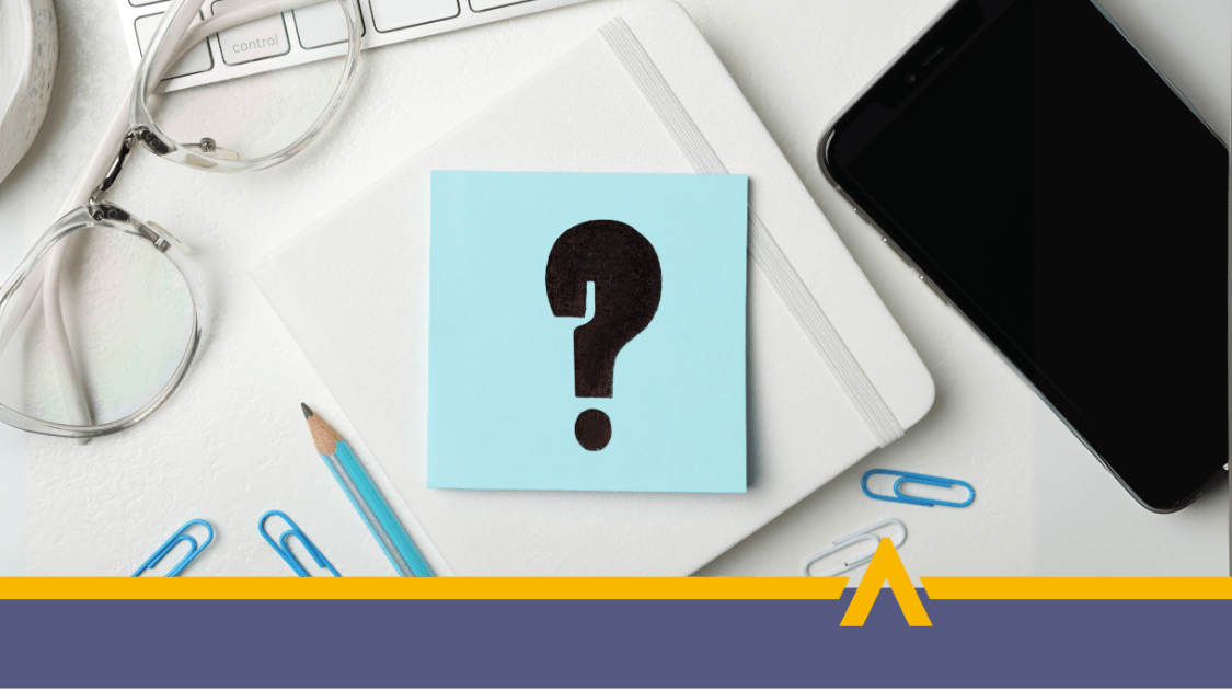 Biurko z notatnikiem, na którym leży niebieska karteczka z dużym znakiem zapytania, obok okulary, ołówek i smartfon.